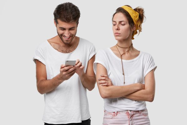 Jealous wife keeps arms folded, looks with displeased expression at husband who ignores her, plays online games on mobile phone, chats with friends, has glad look, isolated over white background